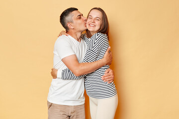 Loving romantic couple man and woman wearing casual clothing standing isolated over beige background hugging aeacj other husband kissing his wife.