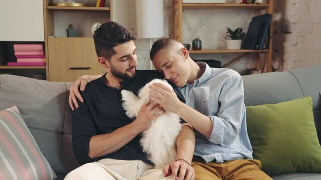 Cute, loving homosexual couple, handsome young men sitting on couch in living room at home and hugging with dog. Showing feelings. Concept of relationship, happiness, animals, lgbt community