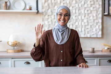 Muslim young woman in hijab sitting in the kitchen at home and smiling talking and greeting to the camera.