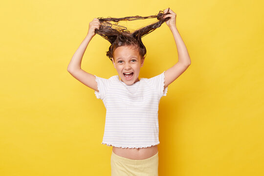 Crazy child girl with shampoo on wet hair wearing white T-shirt standing isolated over yellow background screaming with excitement pulling her hair.