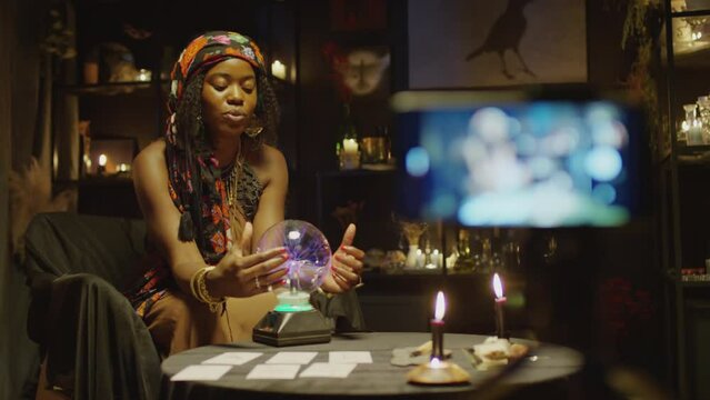 Medium long shot of African American fortune teller touching crystal ball when recording vlog on smartphone