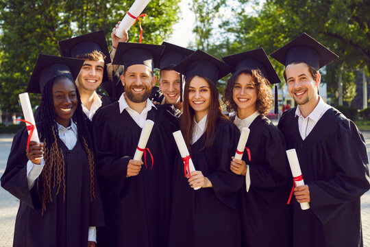 Group of several university students on graduation day. Happy diverse young friends in graduate hats and gowns standing in green campus yard, holding diploma scrolls, looking at camera and smiling