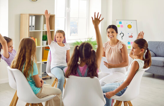Group of school children girls sitting in a circle with female friendly woman psychologist and raising their hands up to ask or answer a question during a meeting on therapy session.