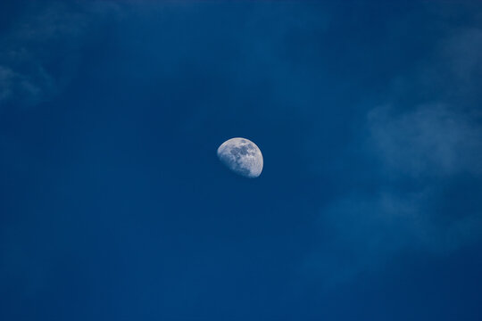 A half moon in a blue sky with puffy clouds. The clouds add a sense of softness and tranquility to the photo. The sky is a deep blue, and it is clear except for the clouds.