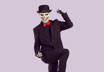 Scary excited man with skeleton makeup celebrating success. Mysterious young man wearing bowler hat, black suit and red bow tie doing winner gesture on isolated studio background