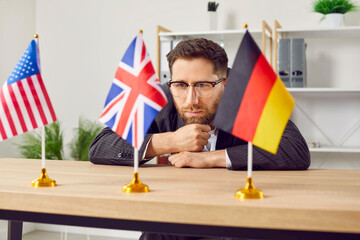 Thoughtful young man sitting at desk in office looking at USA, Great Britain and Germany flags....