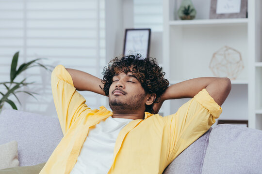 Young man relaxing alone at home on sofa, close-up Hispanic man with hands behind head dozing and dreaming with closed eyes in living room of house