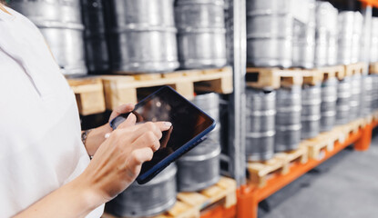 Factory worker with computer tablet inspecting kegs with beer in warehouse of modern brewery stock,...