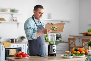 Mature man putting cucumber slices into blender in kitchen