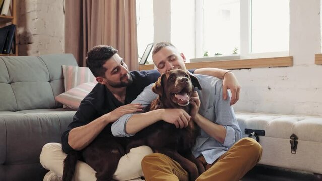 Lovely and relaxed two men, loving couple sitting on couch at home in living room, hugging and petting the dog. Concept of relationship, happiness, animals, lgbt community