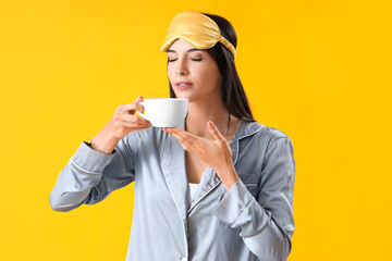 Young woman in pajamas with cup of coffee on yellow background