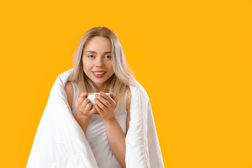 Young woman with blanket and cup of coffee on yellow background