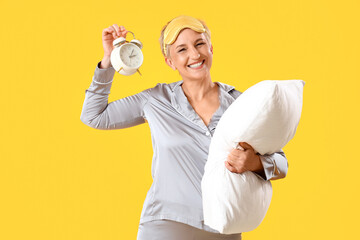 Mature woman in pajamas with pillow and alarm clock on yellow background