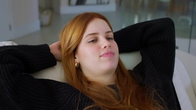 Attractive young woman relaxing indoors leaned on sofa cushions. Woman breath fresh conditioned air folded hands behind head lying on cozy soft couch in living room. Meditation, rest, leisure concept