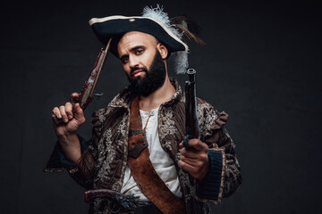Attractive pirate with a black beard, wearing a vest and hat, holding two muskets against a dark...