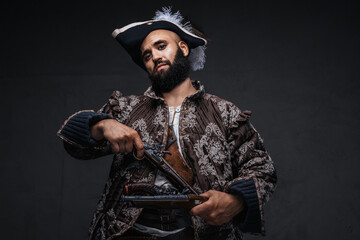 Attractive pirate with a black beard, wearing a vest and hat, holding two muskets against a dark...