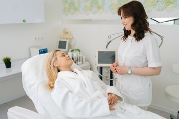 Portrait of smiling female cosmetologist talking with young woman client in white bathrobe lying on couch before rf-lifting procedure in modern light beauty clinic. Concept of non-surgery cosmetology.