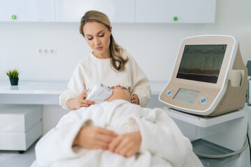 Portrait of concentrated female cosmetologist making ultrasound face lifting massage with professional equipment for young woman lying on couch, in aesthetic medicine clinic with modern interior.