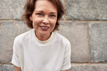 Close-up portrait of attractive mature woman against the background of a stone wall. Middle-aged brunette in a white t-shirt smiles happily. Active lifestyle for the elderly.