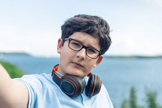 Portrait of eleven years old boy in glasses and wireless headphones, making selfie in front of sea