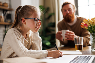 Preadolescent child in eyeglasses writing on notebook near laptop and blurred father in kitchen at home