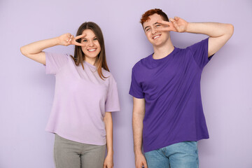 Young couple in t-shirts showing victory gesture on lilac background