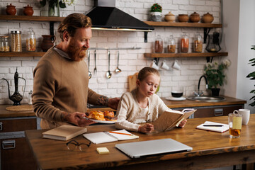 Father holding sweet pastry while daughter with book doing homework in kitchen 