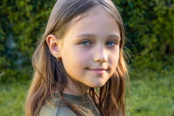 Portrait of a beautiful brunette little teenager girl looking at the camera and smiling, standing on the street against the backdrop of greenery on a summer day. Young curious child raises his eyes