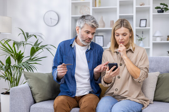 Upset and cheated couple, man and woman disappointed sitting together on couch in living room, using online shopping app on phone, holding bank credit card, refusing money transfer