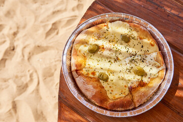 pizza with mozzarella and olives on an aluminum tray on a wooden table on the sand. Fast food to enjoy a summer day at the beach. Copy space