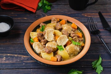 Stewed beef with potatoes, carrots and flour dumplings in a clay plate on a dark wooden background....