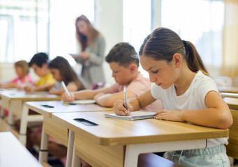 Fototapeta premium Schoolchildren sitting at desks in classroom. Female teacher staning beside desks.