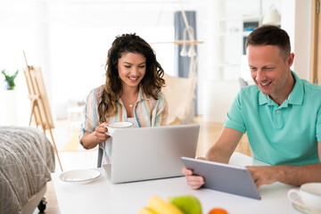 Smiling young couple working from home on their laptop and tablet enjoying morning tea.