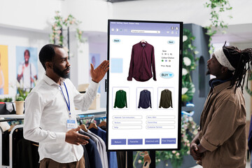 Male assistant showing clothes on interactive kiosk board to young trendy customer in clothing store at mall. Retail employee helping client to choose modern fashion items, self ordering. - Powered by Adobe