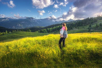 Fototapeta na wymiar Girl on the hill with yellow flowers and green grass in beautiful alpine mountain valley at sunset in summer. Landscape with young woman in alps, rees, sky with clouds. Travel and Hiking. Slovenia