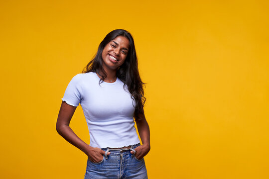 Indian woman posing smiling for photo looking cheerful on camera with hands in pockets. Happy casual female isolated yellow background. Portrait of nice young girl in twenties in positive attitude.