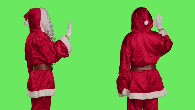 Santa character show palm as no, expressing stop symbol on camera with greenscreen backdrop. Model dressed like saint nick in red costume showing refusal gesture, holiday season concept.