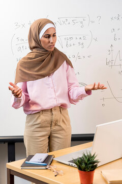 Vertical photo of puzzled Muslim female teacher leading online lesson. Tutor stands against the background of Mathematical board, looks at the laptop, shrugging his hands. Difficulty in learning