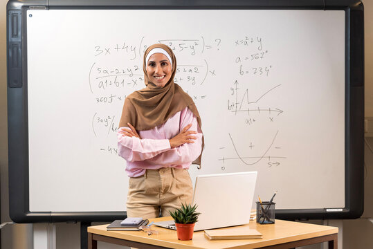 Education concept. Young beautiful Arabic teacher in a hijab stands in the classroom in front of the blackboard with her arms crossed. Confident happy tutor ready to start school year and lesson. 