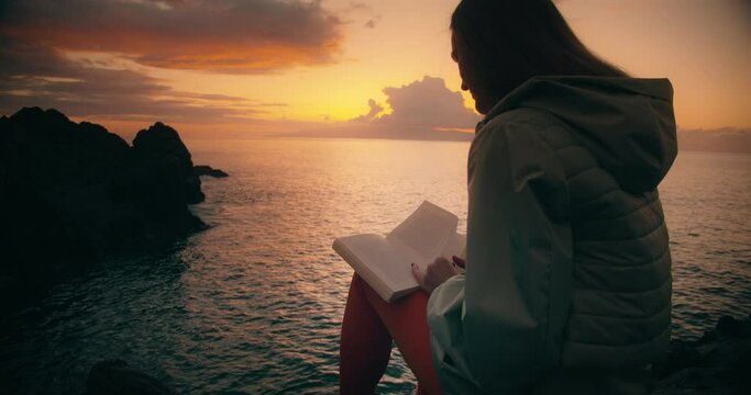 Woman silhouette reading a book on rocky beach with ocean view at sunset.