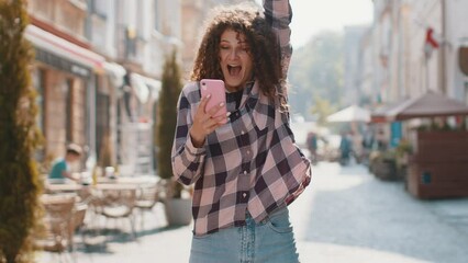 Pretty young woman use mobile smartphone celebrating win good message news, lottery jackpot victory, giveaway online outdoors. Happy girl tourist walking in urban sunshine city street. Town lifestyles