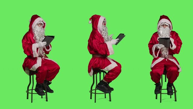 Santa working on digital tablet during christmas eve holiday, expressing december seasonal event. Young man saint nick sitting on chair against full body greenscreen backdrop, using gadget.