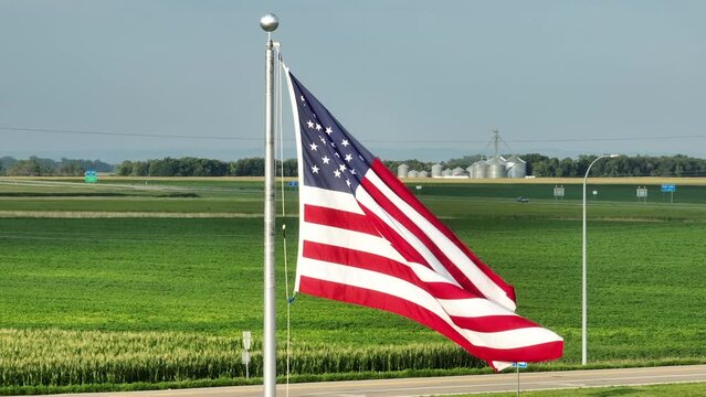 American flag waving in front of corn field and grain elevators in midwest USA. Cinematic aerial establishing shot with long zoom lens.