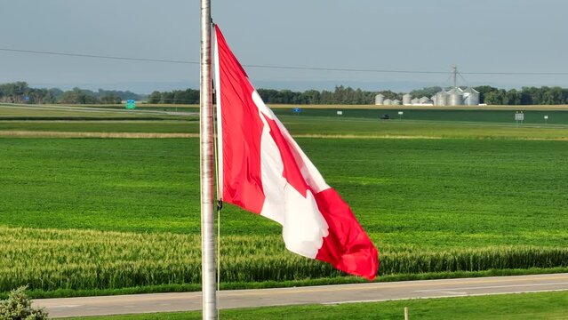 Canadian flag waving with corn fields, farmland, farms, and grain elevators in the distance. Beautiful aerial establishing shot of agriculture production in Canada.
