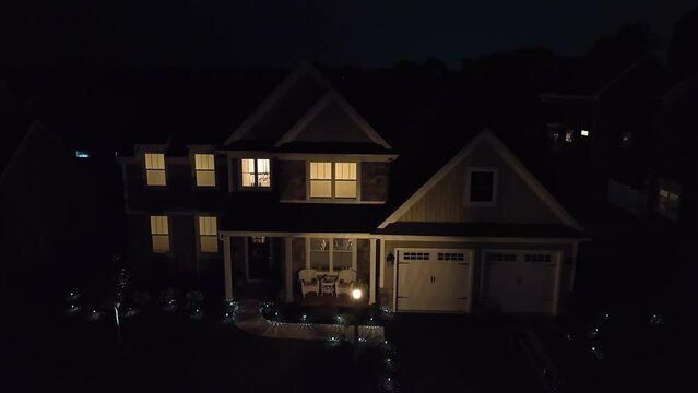 American home with lights on at night. Pitch black shot after dusk. Upscale house in neighborhood. Aerial push in shot.