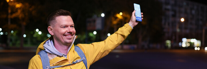 Smiling happy man holding smartphone and meeting friends or call taxi in evening street.