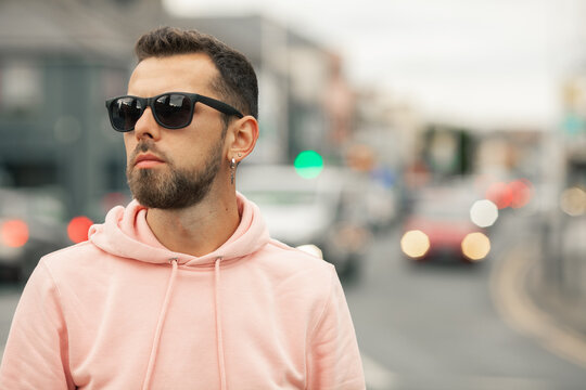 Street fashion concept. Close up portrait of fashionable young man with beard wearing sunglasses, pink sweatshirt, looking somewhere. Text space. Outside shot