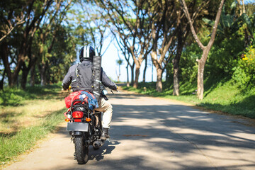 Back view of motorcyclist in black jacket and helmet riding motorbike along country road between tall green trees. 