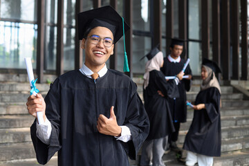 Smiling young Asian male graduate in cap and bachelor gowns showing thumb up and standing against...