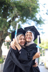 Happy high school Asian muslim girl friend hugging in cap and gown celebrating their graduation